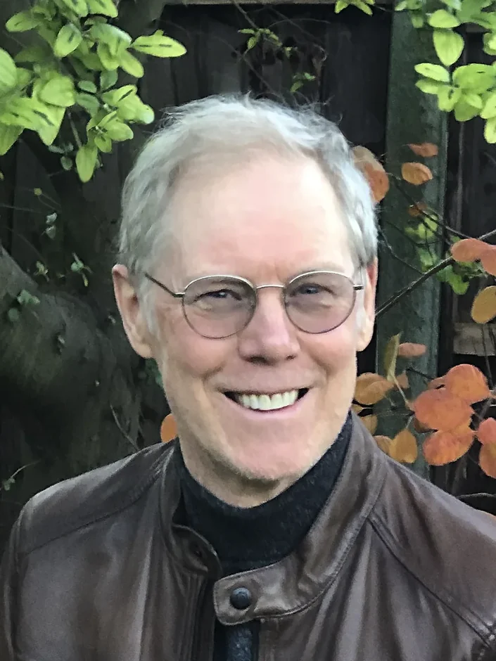 Rob Scotton smiling in round glasses and a brown leather jacket, with autumn leaves behind him